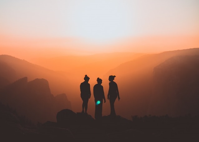 Three women stand in sillhoute atop a mountain on a hazy yellow day at sunset.